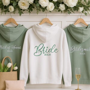 May include: Three hooded sweatshirts on wooden hangers. One is white with "Bride" in green script, the date 28.6.24. The other two are sage green, one with "Maid of Honour" and the other with "Bridesmaid", both with the date 28.6.24.