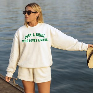 May include: Cream-coloured sweatshirt and shorts set with the text "JUST A BRIDE WHO LOVES A MARG." in green. The person is wearing black sunglasses and holding a pair of flip-flops. The background is a body of water.