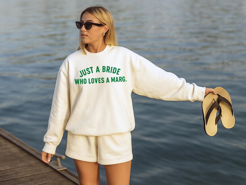 May include: Cream-coloured sweatshirt and shorts set with the text "JUST A BRIDE WHO LOVES A MARG." in green. The person is wearing black sunglasses and holding a pair of flip-flops. The background is a body of water.