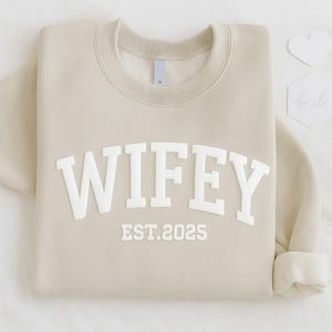 May include: A beige sweatshirt featuring the word "WIFEY" in large, white, raised letters, with "EST. 2025" below. The sweatshirt is folded with a rolled cuff. A small white heart and a card that says "bride" are in the background.
