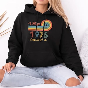 Könnte beinhalten: Schwarzer Kapuzenpullover mit Retro-Design. Die Grafik zeigt das Wort "Vintage" über dem Jahr "1976" und die Worte "Original Parts" in einem farbenfrohen, geschichteten Design. Der Pullover hat eine Vordertasche.