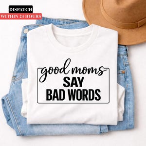 May include: White long-sleeved t-shirt with the text "good moms SAY BAD WORDS" in a black, handwritten-style font. The shirt is folded on top of blue jeans. A brown hat is visible in the background.