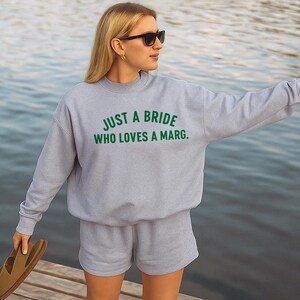 May include: A light grey sweatshirt and shorts set with the text "JUST A BRIDE WHO LOVES A MARG." in green. The person is wearing sunglasses and holding sandals. The background is a body of water.