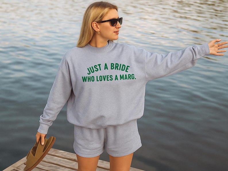 May include: A light grey sweatshirt and shorts set with the text "JUST A BRIDE WHO LOVES A MARG." in green. The person is wearing sunglasses and holding sandals. The background is a body of water.