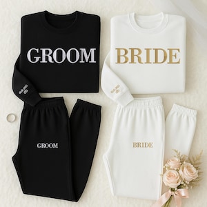 May include: Matching black and white wedding day loungewear sets. The black set includes a sweatshirt with "GROOM" in white lettering and matching sweatpants. The white set has "BRIDE" in gold lettering and matching sweatpants. Both sets have the wedding date on the sleeve.