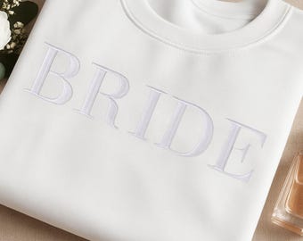 May include: A white crewneck sweatshirt with the word "BRIDE" embroidered in white. The sweatshirt is made of a soft material and is perfect for a wedding or bridal shower. The design is simple and elegant.