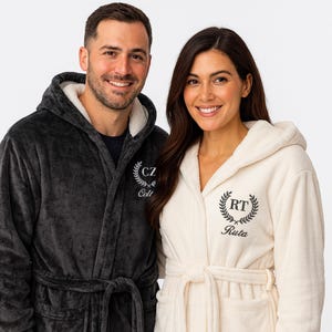 Op de afbeelding: Twee mensen dragen bijpassende badjassen met capuchon. Eén is antracietgrijs, de andere is crèmekleurig. Elke badjas heeft een gepersonaliseerd geborduurd ontwerp met initialen en een naam. De badjassen hebben een zachte, pluche textuur en een strikriem.