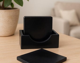 Puede incluir: Un juego de cuatro posavasos cuadrados negros con un soporte a juego. Un posavasos se coloca fuera del soporte sobre una superficie de madera clara. Los posavasos y el soporte están hechos de un material oscuro y sólido.