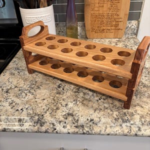 May include: A two-tiered wooden egg holder with 20 circular openings. The holder has a natural wood finish and features handles on each end. The item is displayed on a speckled countertop.