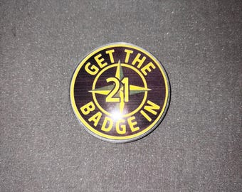Könnte beinhalten: Ein runder Anstecker mit dem Text "GET THE BADGE IN 21" in gelber Schrift. Der Anstecker hat einen schwarzen und braunen Hintergrund mit einem gelben Kompassdesign in der Mitte. Der Anstecker hat einen gelben Rand.