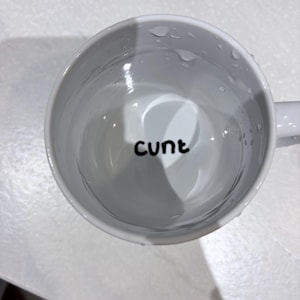 K&ouml;nnte beinhalten: Ein wei&szlig;er Keramikbecher, gef&uuml;llt mit einer klaren Fl&uuml;ssigkeit, mit dem Wort "cunt" in schwarzen, kleingeschriebenen, serifenlosen Buchstaben am Boden des Innenraums. Der Becher hat eine glatte, gl&auml;nzende Oberfl&auml;che mit sichtbaren Wassertropfen.