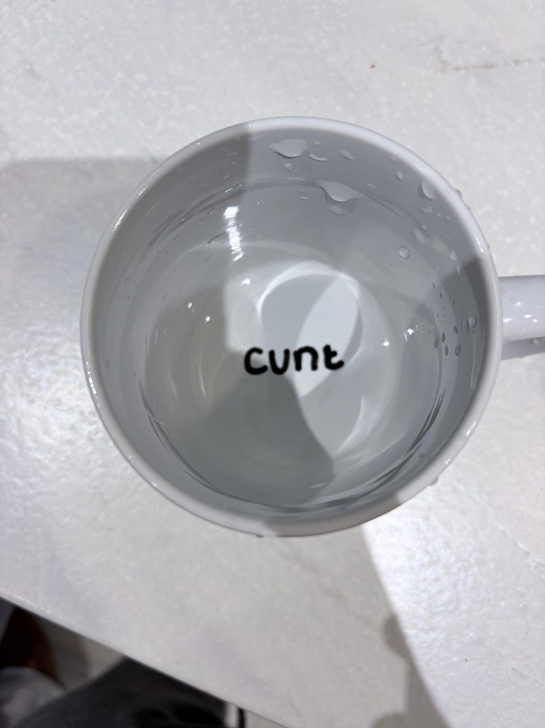 K&ouml;nnte beinhalten: Ein wei&szlig;er Keramikbecher, gef&uuml;llt mit einer klaren Fl&uuml;ssigkeit, mit dem Wort "cunt" in schwarzen, kleingeschriebenen, serifenlosen Buchstaben am Boden des Innenraums. Der Becher hat eine glatte, gl&auml;nzende Oberfl&auml;che mit sichtbaren Wassertropfen.