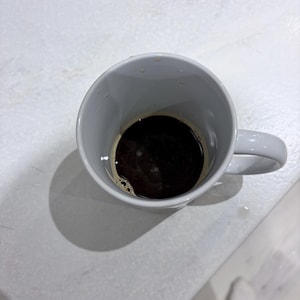 K&ouml;nnte beinhalten: Eine wei&szlig;e Keramiktasse gef&uuml;llt mit schwarzem Kaffee. Die Tasse hat eine glatte, gl&auml;nzende Oberfl&auml;che und einen geschwungenen Henkel auf der rechten Seite. Die dunkle Fl&uuml;ssigkeit im Inneren zeigt eine d&uuml;nne Crema-Schicht, die auf frisch gebr&uuml;hten Kaffee hindeutet.