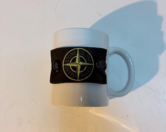 Könnte beinhalten: Weißer Keramikbecher mit einem schwarzen Band mit einem gold-schwarzen Logo. Der Becher hat einen gebogenen Griff und steht auf einer weißen Oberfläche.