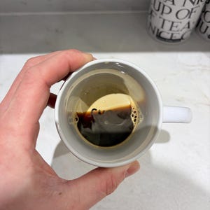 K&ouml;nnte beinhalten: Ein wei&szlig;er Keramikbecher mit einer dunklen Fl&uuml;ssigkeit und einer hellbraunen Schaumschicht. Im Inneren des Bechers ist der schwarze Schriftzug "Cup" in der N&auml;he des Fl&uuml;ssigkeitsspiegels aufgedruckt. Der Becher hat einen einfachen Henkel und eine glatte, wei&szlig;e Oberfl&auml;che.