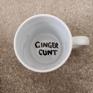 K&ouml;nnte beinhalten: Eine wei&szlig;e Keramiktasse mit Henkel, direkt von oben betrachtet. Auf dem inneren Boden der Tasse befindet sich der Text 'GINGER CUNT', der in einer fetten, schwarzen, handgeschriebenen Schriftart auf dem wei&szlig;en Untergrund steht.