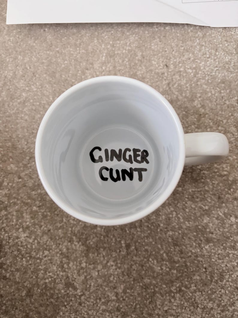 K&ouml;nnte beinhalten: Eine wei&szlig;e Keramiktasse mit Henkel, direkt von oben betrachtet. Auf dem inneren Boden der Tasse befindet sich der Text 'GINGER CUNT', der in einer fetten, schwarzen, handgeschriebenen Schriftart auf dem wei&szlig;en Untergrund steht.