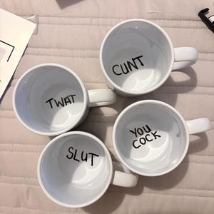 K&ouml;nnte beinhalten: Vier wei&szlig;e Keramikbecher von oben betrachtet, jeder mit schwarzem Text auf dem inneren Boden. Die Becher zeigen die W&ouml;rter CUNT, TWAT, SLUT und YOU COCK in einer einfachen serifenlosen Schrift und verf&uuml;gen &uuml;ber standardm&auml;&szlig;ige Henkel.