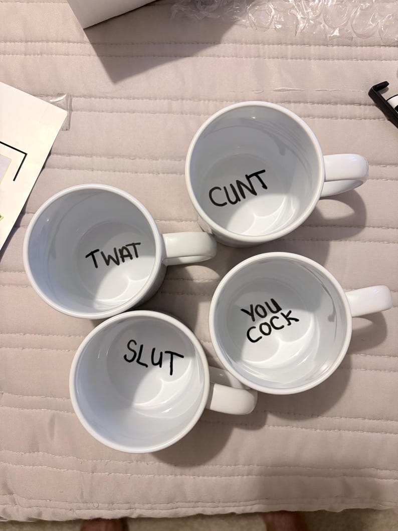 K&ouml;nnte beinhalten: Vier wei&szlig;e Keramikbecher von oben betrachtet, jeder mit schwarzem Text auf dem inneren Boden. Die Becher zeigen die W&ouml;rter CUNT, TWAT, SLUT und YOU COCK in einer einfachen serifenlosen Schrift und verf&uuml;gen &uuml;ber standardm&auml;&szlig;ige Henkel.