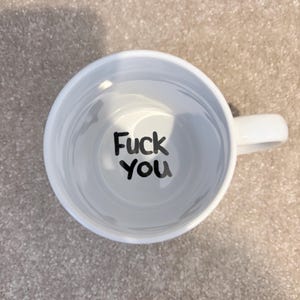 K&ouml;nnte beinhalten: Eine wei&szlig;e Keramiktasse, von direkt oben betrachtet, mit einem Henkel auf der rechten Seite. Auf dem inneren Boden der Tasse ist der Text "Fuck you" in einer fetten, schwarzen, handgeschriebenen Schriftart vor dem wei&szlig;en Hintergrund gedruckt.