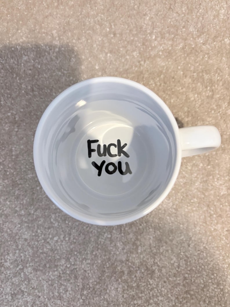 K&ouml;nnte beinhalten: Eine wei&szlig;e Keramiktasse, von direkt oben betrachtet, mit einem Henkel auf der rechten Seite. Auf dem inneren Boden der Tasse ist der Text "Fuck you" in einer fetten, schwarzen, handgeschriebenen Schriftart vor dem wei&szlig;en Hintergrund gedruckt.