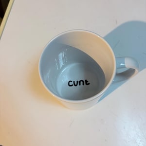 K&ouml;nnte beinhalten: Ein wei&szlig;er Keramikbecher mit Henkel, der das Wort "cunt" in schwarzer, kleingeschriebener, serifenloser Schrift auf dem inneren Boden des Bechers aufgedruckt hat. Der Becher ist aus der Vogelperspektive zu sehen, die die &Ouml;ffnung und den Text zeigt.