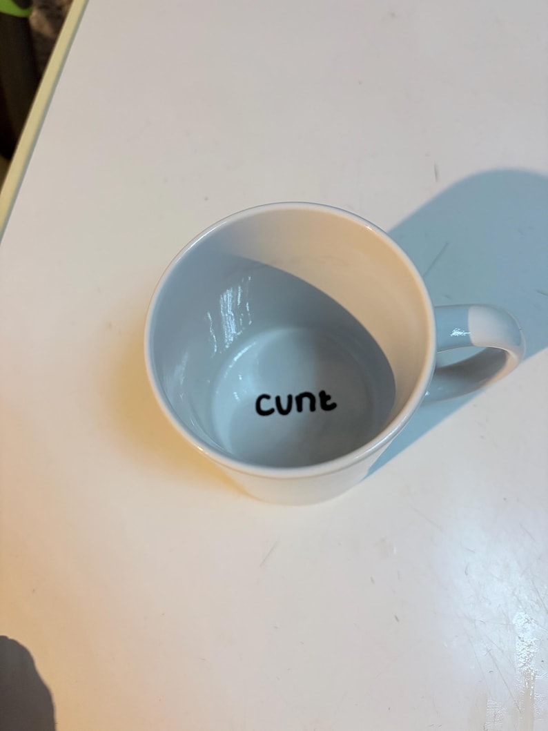 K&ouml;nnte beinhalten: Ein wei&szlig;er Keramikbecher mit Henkel, der das Wort "cunt" in schwarzer, kleingeschriebener, serifenloser Schrift auf dem inneren Boden des Bechers aufgedruckt hat. Der Becher ist aus der Vogelperspektive zu sehen, die die &Ouml;ffnung und den Text zeigt.