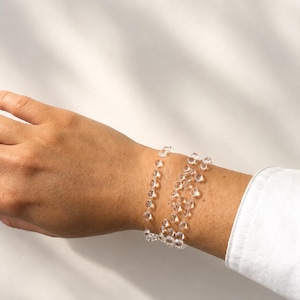 May include: A multi-strand bracelet composed of small, clear, spherical beads strung on a thin, transparent cord. The beads are arranged in multiple loops around the wrist, creating a layered, delicate appearance with a reflective, glass-like quality.