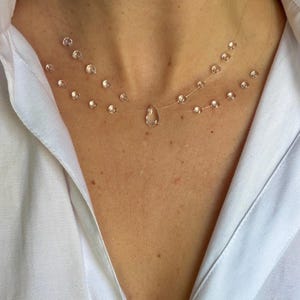Può includere: Una collana a doppio filo con perline rotonde e trasparenti infilate su un filo sottile e invisibile. Il design presenta un ciondolo centrale a forma di goccia appeso al filo inferiore, creando un effetto fluttuante sulla pelle.