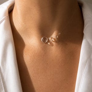 Könnte beinhalten: Eine transparente Halskette mit vier kugelförmigen Perlen unterschiedlicher Größe, die auf einer dünnen, durchsichtigen Nylonschnur aufgereiht sind. Die Perlen sind horizontal angeordnet, wobei die größte Kugel links und drei kleiner werdende Kugeln rechts liegen.