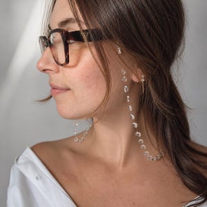 Op de afbeelding: Een brilketting met kleine, doorzichtige, ronde kralen geregen aan een dun, flexibel koord. De ketting is bevestigd aan de pootjes van een donkerbruine bril met schildpadpatroon en hangt langs de zijkanten van het montuur naar beneden.
