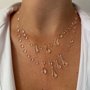May include: A clear, layered necklace featuring small, round beads and teardrop-shaped pendants. The necklace is worn against a white shirt, creating a minimalist and elegant aesthetic. The design is delicate and appears to float on the skin.