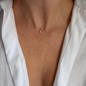 Könnte beinhalten: Eine zarte Halskette mit Tropfenanhänger. Der Anhänger ist ein kleiner, klarer, tropfenförmiger Anhänger, der an einer fast unsichtbaren, dünnen, klaren Schnur befestigt ist. Die Halskette wird über einem weißen Hemd getragen.