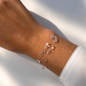 Op de afbeelding: Een delicate armband met meerdere kleine, heldere, ronde kralen geregen aan een dun, transparant draadje. De kralen zijn verspreid over het koord, wat een zwevend effect op de huid creëert. Het ontwerp is eenvoudig en licht van gewicht.