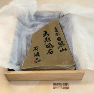 May include: A tan, triangular whetstone with Japanese characters, nestled in a wooden box and wrapped in white tissue paper. The text on the stone includes "Kyoto" and "Natural Whetstone". The bottom of the box reads "DAIKUDOU WHETSTONE".