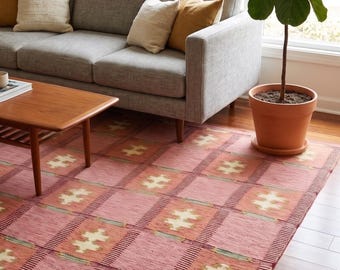 Puede incluir: Una alfombra estampada en tonos rosas y naranjas con un diseño geométrico. La alfombra está colocada sobre un suelo de madera en un salón, con un sofá gris y una planta en maceta en el fondo.