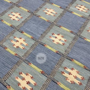May include: A woven rug featuring a grid pattern of alternating light blue and sage green squares. Each square contains a cream-coloured geometric motif with red accents. The grid is separated by thin black and white striped lines. Text reads RUG MANIA CREATIONS.