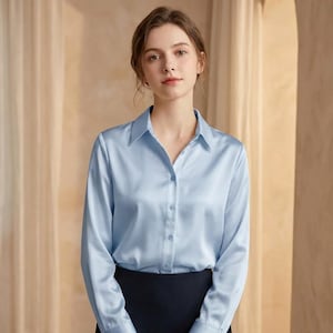 May include: A light blue button-down shirt with long sleeves. The shirt has a classic collar and is made of a silky, smooth fabric. The model is wearing a black skirt. The shirt is a solid color and has a subtle sheen.