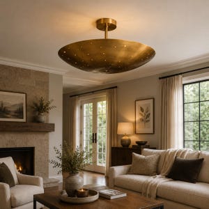 Könnte beinhalten: Eine messingfarbene, schalenförmige Deckenleuchte mit kleinen Perforationen. Das Licht wird von einer zentralen Stange abgehängt und beleuchtet ein Wohnzimmer mit neutralen Tönen, einem Kamin und einem Sofa.
