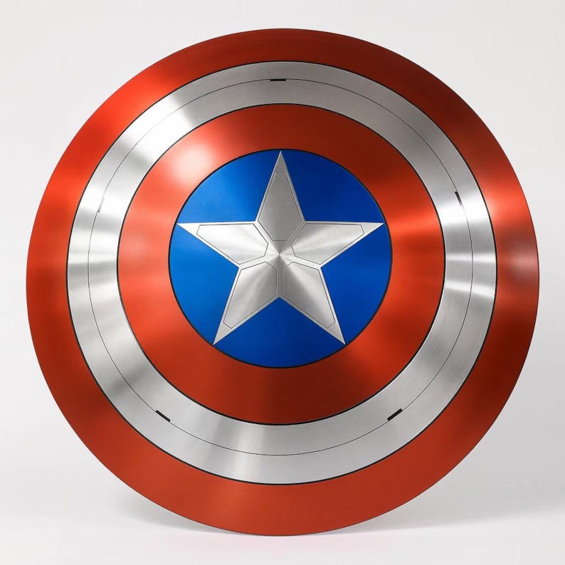 Op de afbeelding: Een rond schild met een rode buitenring, een zilveren middenring en een blauw centrum. In het midden staat een grote, zilveren vijfpuntige ster. Het schild heeft een metallic afwerking en is waarschijnlijk een rekwisiet of verzamelobject.
