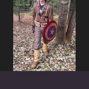 Op de afbeelding: Een persoon in een Captain America-kostuum, met een schild van 56 cm. Het kostuum omvat een bruine jas, broek, laarzen en een helm. Het schild is rood, wit en blauw met een ster in het midden. De persoon staat in een bosrijk gebied.
