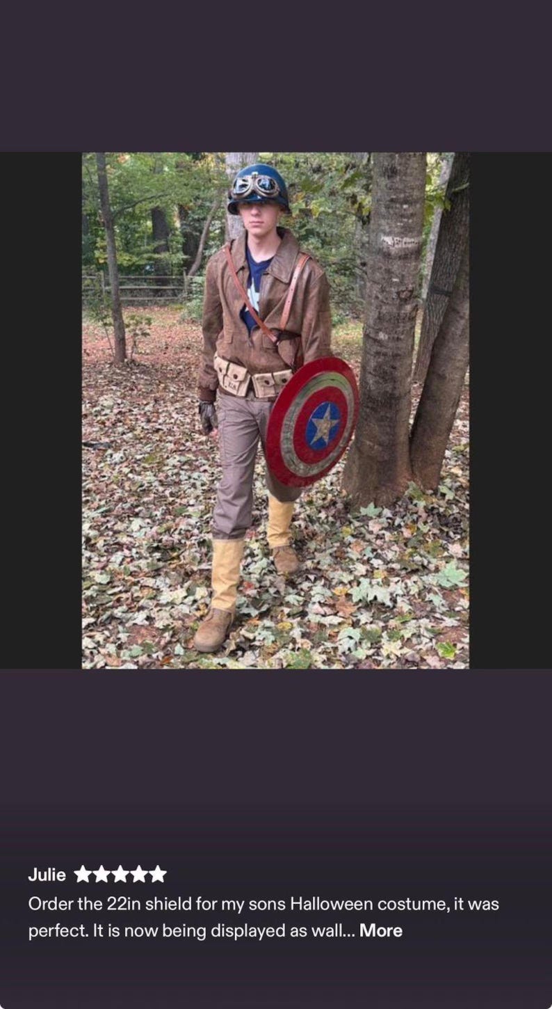 Op de afbeelding: Een persoon in een Captain America-kostuum, met een schild van 56 cm. Het kostuum omvat een bruine jas, broek, laarzen en een helm. Het schild is rood, wit en blauw met een ster in het midden. De persoon staat in een bosrijk gebied.
