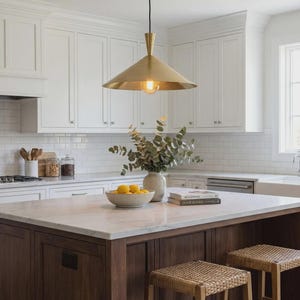 Könnte beinhalten: Helle, moderne Küche mit weißen Schränken, einer Insel mit Marmorplatte und einer Pendelleuchte aus Messing. Die Insel hat einen dunklen Holzsockel und zwei gewebte Hocker. Eine Schale mit Zitronen und eine Vase mit Grün.
