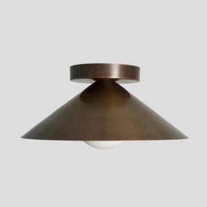 Könnte beinhalten: Eine moderne, bündig montierte Deckenleuchte in Bronzefarbe. Die Leuchte hat einen konischen Schirm und einen runden Sockel. Eine weiße, kugelförmige Glühbirne ist am unteren Ende des Schirms sichtbar. Die Leuchte ist für den Innenbereich konzipiert.