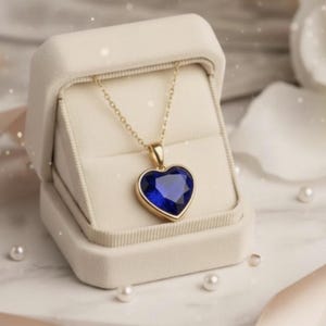 May include: A gold necklace featuring a heart-shaped, deep blue pendant, presented in an open, cream-coloured jewellery box. The pendant is approximately 2.5 cm wide. The box rests on a marble surface, with scattered pearls and a satin ribbon.
