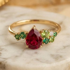 Peut inclure: Une bague en or ornée d'une pierre rouge centrale en forme de poire, entourée d'un groupe de petites pierres vertes rondes et d'une pierre blanche transparente de chaque côté, montée sur un anneau fin en or poli avec un sertissage délicat.