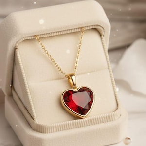 May include: A gold necklace with a heart-shaped, red gemstone pendant displayed in an open, cream-colored jewelry box. The necklace is on a marble surface with scattered pearls and a satin ribbon.