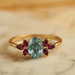 Può includere: Un anello d'oro con una pietra preziosa ovale azzurra al centro, circondata da pietre preziose rosso scuro più piccole, a formare un motivo floreale. L'anello è appoggiato su una superficie di marmo.
