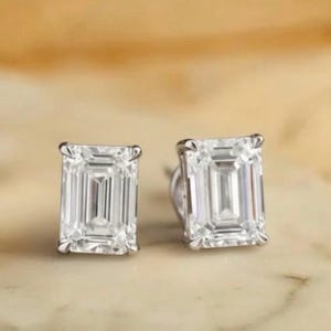 May include: A pair of square-cut, clear gemstone stud earrings. The earrings are set in a silver metal and have a rectangular shape. The background is a light brown and white marble surface.