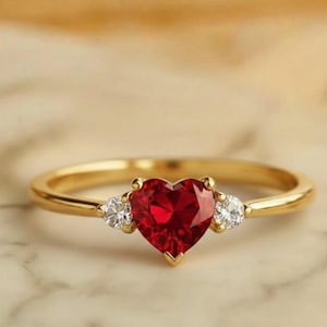 May include: A gold ring with a heart-shaped, red gemstone center and two smaller white gemstones on either side. The ring is set on a marble surface. The text "GARDEN MINIMALIST GARD" is at the top.